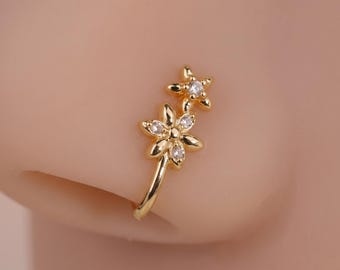 May include: A gold-toned nose cuff featuring two flower-shaped motifs arranged vertically. The larger bottom flower has five petals with a central clear stone, while the smaller top flower has five petals with a single clear stone.