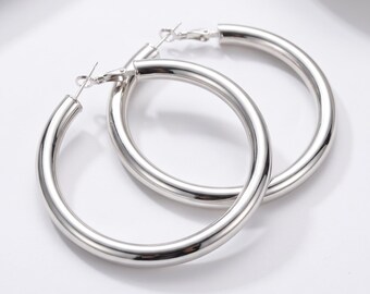 May include: A pair of large, polished silver-tone hoop earrings with a smooth, rounded tubular design. Each earring features a hinged post closure mechanism at the top, showcasing a reflective metallic finish and a classic circular silhouette.