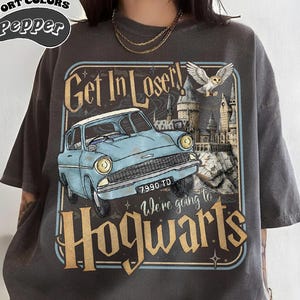 May include: A charcoal gray short-sleeve t-shirt featuring a graphic print of a light blue vintage car with the license plate 7990 TD, a castle, and an owl. The text reads Get In Loser! We're going to Hogwarts. The top left corner says Comfort Colors Pepper.