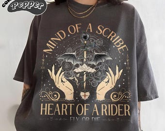 May include: A charcoal gray short-sleeve t-shirt featuring a graphic print with the text "MIND OF A SCRIBE HEART OF A RIDER FLY OR DIE". The design includes two hands, a central sword with wings, celestial symbols, and a "COMFORT COLORS Pepper" logo.