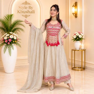May include: A beige floor-length anarkali dress with long sleeves, featuring a vibrant red embroidered bodice with mirror work and hanging tassels. The dress includes a matching beige dupatta and a red embroidered border at the hemline.
