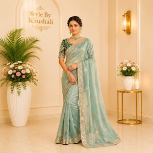 May include: A light blue saree with a dark green embroidered blouse. The saree features silver embellishments and a decorative border. The ensemble includes a necklace and earrings, with the text Style By Khushali visible on the wall.