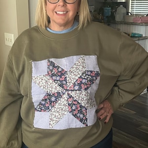 Puede incluir: Una sudadera de cuello redondo color verde oliva con una gran aplicación cuadrada de bloque de edredón en la parte delantera. La aplicación muestra un patrón de estrella construido con tela azul claro y tela con estampado floral en tonos azules y rosas.