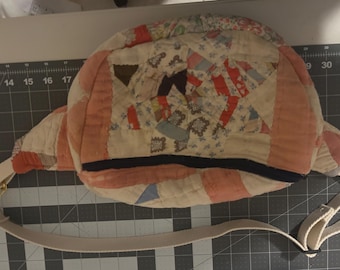 Puede incluir: Una riñonera confeccionada con tela de patchwork acolchada que presenta varios patrones florales y geométricos en tonos crema, melocotón y rojo. El bolso incluye un cierre de cremallera horizontal negro y una correa ajustable de color blanco roto.