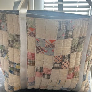 May include: A tote bag constructed from a quilted patchwork fabric featuring a variety of small square patterns in muted tones of white, blue, red, and brown. The bag includes two long, smooth, light gray straps attached to the exterior front.
