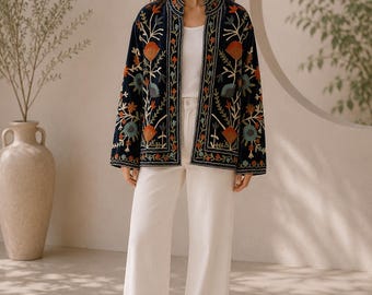 May include: A dark navy blue open-front jacket featuring intricate multi-colored floral embroidery in shades of orange, teal, and cream. The jacket has long sleeves, a mandarin collar, and is worn over a plain white tank top and white wide-leg trousers.