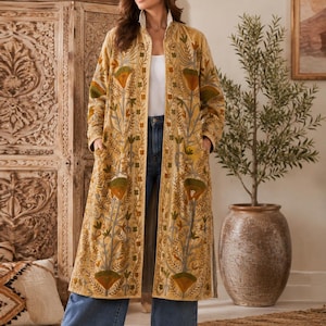 Può includere: Un lungo cappotto color giallo senape con apertura frontale, caratterizzato da intricati ricami floreali nei toni del verde, dell'arancione e del grigio. Il capo presenta maniche lunghe, tasche laterali e orlo dritto, indossato su un top bianco e jeans blu.