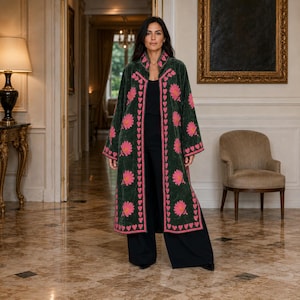 Pode incluir: Um casaco comprido de veludo verde-escuro com um padrão floral bordado em rosa vibrante e uma borda de corações repetidos ao longo da abertura frontal e dos punhos. O casaco é usado sobre um top preto simples e calças pretas largas.