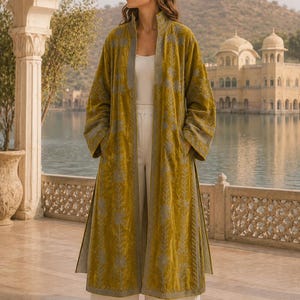Puede incluir: Un abrigo largo tipo duster de terciopelo verde oliva con cuello alto, mangas largas y un intrincado bordado floral gris en los paneles frontales y los puños. La prenda se lleva sobre una camiseta de tirantes blanca y pantalones blancos anchos.