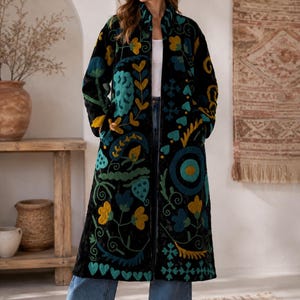 Pode incluir: Um casaco comprido preto com bordados florais e abstratos multicoloridos em tons de azul-petróleo, amarelo-mostarda e azul-claro. O casaco tem gola alta, mangas compridas e uma silhueta reta, usado sobre um top branco e calças de ganga azuis.