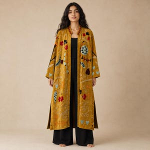 Puede incluir: Un abrigo largo de color amarillo mostaza con frente abierto y bordados florales y de hojas multicolores en tonos rojo, azul y verde. La prenda tiene mangas largas, cuello tipo mandarín y aberturas laterales, sobre un top y pantalones negros.
