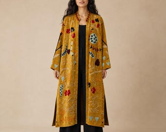 May include: A long, mustard-yellow open-front coat featuring intricate, multi-colored floral and leaf embroidery in shades of red, blue, and green. The garment has long sleeves, a mandarin-style collar, and side slits, worn over a black top and pants.