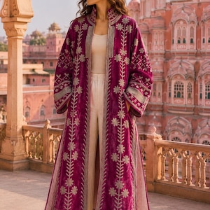 May include: A long, magenta velvet duster coat featuring intricate silver floral embroidery along the front panels and sleeves. The coat is worn over a cream-coloured tank top and wide-leg trousers, showcasing a high collar and open front design.