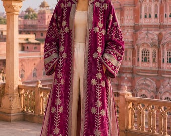 May include: A long, magenta velvet duster coat featuring intricate silver floral embroidery along the front panels and sleeves. The coat is worn over a cream-colored tank top and wide-leg trousers, showcasing a high collar and open front design.