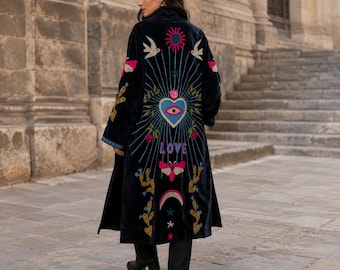 May include: A black velvet duster coat featuring intricate colorful embroidery on the back. The design includes a central heart with an eye, the word LOVE, a sun, birds, moths, cacti, and a crescent moon in shades of pink, blue, yellow, and white.