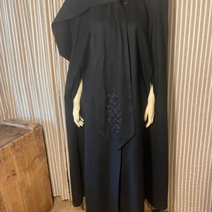 May include: A floor-length black cape featuring a high collar, a single button closure at the neck, and a long, draped front panel. The front panel is decorated with tonal black embroidery in a scrolling, ornate pattern.