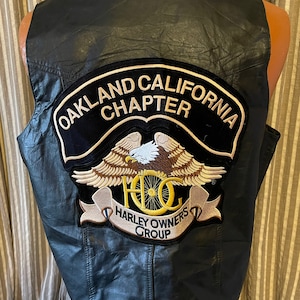May include: Black leather vest with an embroidered Harley Owners Group (HOG) patch. The patch features an eagle with wings spread, a motorcycle wheel, and the text "Harley Owners Group". The vest also has a patch that says "Oakland California Chapter".