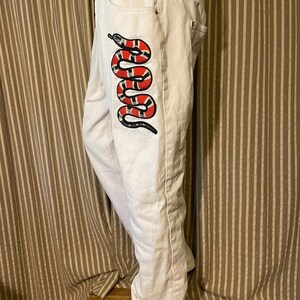 May include: White denim jeans with a red, black, and white snake embroidered on the side seam.