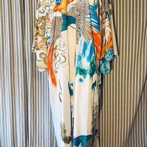 May include: A long, flowing kimono-style robe with a V-neck. The robe features a cream-colored background with a vibrant print of orange, teal, blue, and gold abstract figures and patterns. The sleeves are wide and the robe reaches the floor.