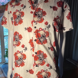 May include: A short-sleeved button-down shirt in an off-white color featuring a repeating pattern of human skulls, vibrant red roses, and small black and white snakes. The shirt has a standard collar and dark buttons down the front center.