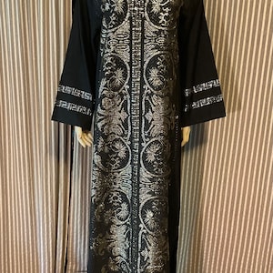 May include: Black floor-length dress with silver metallic embroidery. The dress features a V-neck, long sleeves, and intricate floral and geometric patterns. The sleeves and neckline are trimmed with silver accents. The dress is displayed on a mannequin.