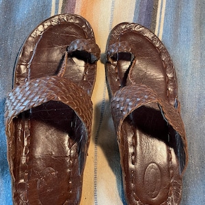 May include: A pair of dark brown leather sandals featuring a toe loop design and a wide, woven leather strap across the top. The footbed is smooth with visible stitching along the edges and an oval-shaped indentation near the heel area.
