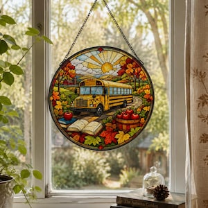 May include: A circular stained glass suncatcher featuring a yellow school bus on a road, surrounded by autumn foliage, a basket of red apples, and an open book. The piece is suspended by a metal chain against a window with a view of trees.