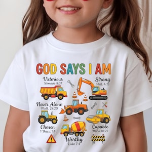 Peut inclure: Un t-shirt blanc arborant un graphique coloré de véhicules de chantier, dont un camion-benne, une pelleteuse, un tracteur et une bétonnière, avec le texte "GOD SAYS I AM" et diverses phrases inspirantes et références bibliques.