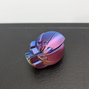 Op de afbeelding: Een klein, driedimensionaal object met een afgeronde, gesegmenteerde schaal met een metallic kleurverloop dat verschuift tussen tinten diepblauw, paars en magenta. Het oppervlak vertoont een fijne, gelaagde textuur van 3D-printen.