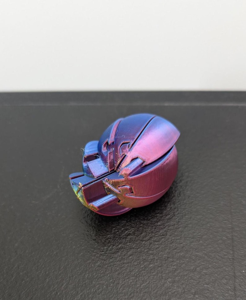 Op de afbeelding: Een klein, driedimensionaal object met een afgeronde, gesegmenteerde schaal met een metallic kleurverloop dat verschuift tussen tinten diepblauw, paars en magenta. Het oppervlak vertoont een fijne, gelaagde textuur van 3D-printen.
