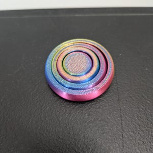 Peut inclure: Un fidget spinner circulaire arborant un d&eacute;grad&eacute; arc-en-ciel vibrant avec des teintes m&eacute;talliques bleues, vertes, jaunes et roses. L'objet pr&eacute;sente un capuchon central textur&eacute; et des d&eacute;tails en anneaux concentriques sur sa surface plate.