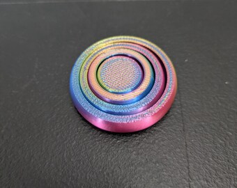 Może przedstawiać: Okrągły fidget spinner z żywym gradientem tęczy w metalicznych odcieniach niebieskiego, zielonego, żółtego i różowego. Urządzenie posiada teksturowaną, radełkowaną nasadkę środkową oraz detale w formie koncentrycznych pierścieni na powierzchni.