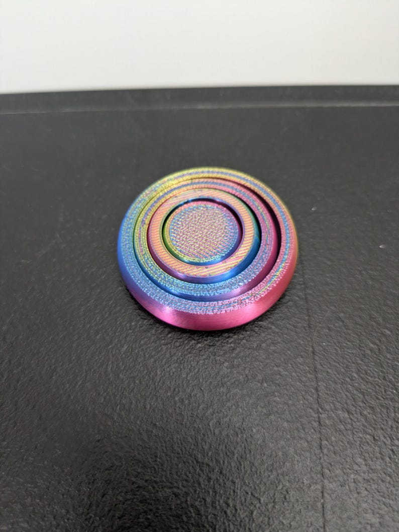Peut inclure: Un fidget spinner circulaire arborant un d&eacute;grad&eacute; arc-en-ciel vibrant avec des teintes m&eacute;talliques bleues, vertes, jaunes et roses. L'objet pr&eacute;sente un capuchon central textur&eacute; et des d&eacute;tails en anneaux concentriques sur sa surface plate.