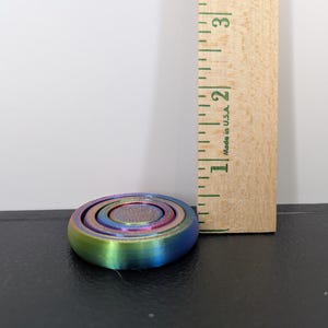 Peut inclure: Un fidget spinner circulaire aux couleurs arc-en-ciel iris&eacute;es avec un centre textur&eacute;, plac&eacute; &agrave; c&ocirc;t&eacute; d'une r&egrave;gle en bois montrant un diam&egrave;tre d'environ 3,8 cm. L'objet pr&eacute;sente des anneaux concentriques et une finition m&eacute;tallique.