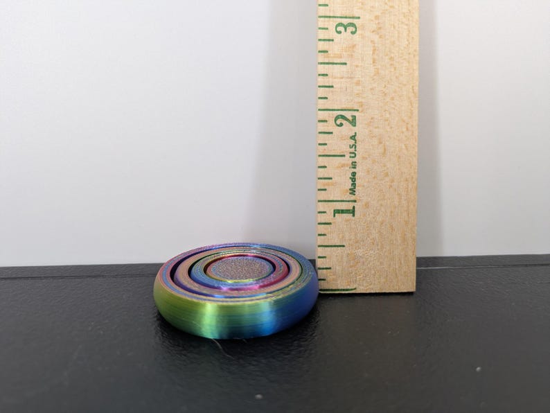 Peut inclure: Un fidget spinner circulaire aux couleurs arc-en-ciel iris&eacute;es avec un centre textur&eacute;, plac&eacute; &agrave; c&ocirc;t&eacute; d'une r&egrave;gle en bois montrant un diam&egrave;tre d'environ 3,8 cm. L'objet pr&eacute;sente des anneaux concentriques et une finition m&eacute;tallique.
