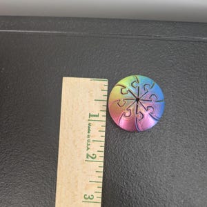Op de afbeelding: Een ronde, meerkleurige knoop met kleurverloop en een puzzelstukjespatroon, geplaatst naast een houten liniaal die een diameter van ongeveer 3,2 centimeter aangeeft. De knoop vertoont een spectrum van geel, roze en blauw.