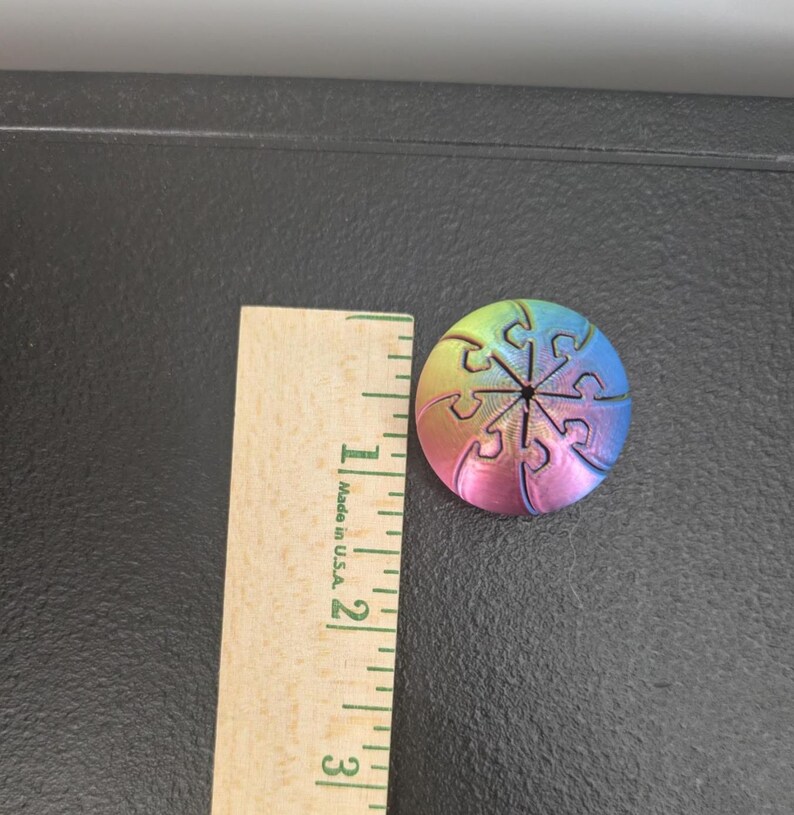 Op de afbeelding: Een ronde, meerkleurige knoop met kleurverloop en een puzzelstukjespatroon, geplaatst naast een houten liniaal die een diameter van ongeveer 3,2 centimeter aangeeft. De knoop vertoont een spectrum van geel, roze en blauw.