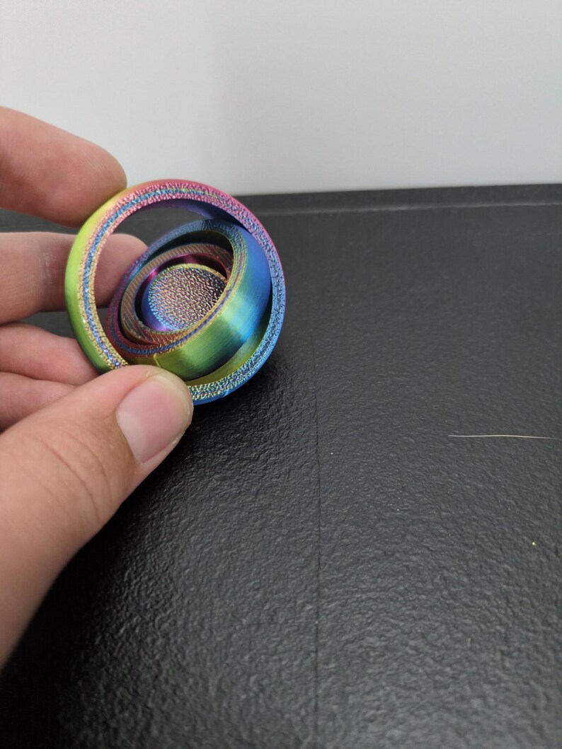 Peut inclure: Un jouet anti-stress iridescent multicolore compos&eacute; d'une s&eacute;rie d'anneaux concentriques imbriqu&eacute;s avec une finition m&eacute;tallique textur&eacute;e, passant par des nuances de vert, bleu, violet et rose dans une disposition circulaire et sph&eacute;rique.