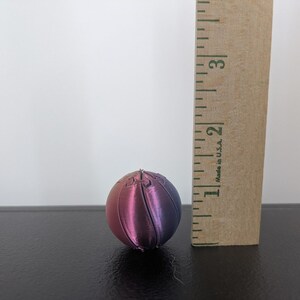 Op de afbeelding: Een bolvormig object met een kleurverloop van roze naar paars, voorzien van verticale gesegmenteerde lijnen. Een houten liniaal staat verticaal naast de bol en geeft een hoogte aan van ongeveer 3,2 centimeter.