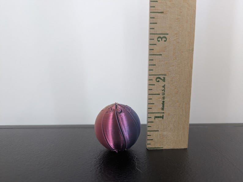 Op de afbeelding: Een bolvormig object met een kleurverloop van roze naar paars, voorzien van verticale gesegmenteerde lijnen. Een houten liniaal staat verticaal naast de bol en geeft een hoogte aan van ongeveer 3,2 centimeter.