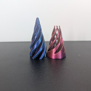 Może przedstawiać: Dwa stożkowe obiekty o spiralnej konstrukcji z otwartą ramą. Wyższy obiekt ma metaliczne wykończenie z gradientem niebiesko-fioletowym, a kr&oacute;tszy obiekt ma metaliczne wykończenie z gradientem r&oacute;żowo-fioletowym z widocznymi pionowymi żebrami.