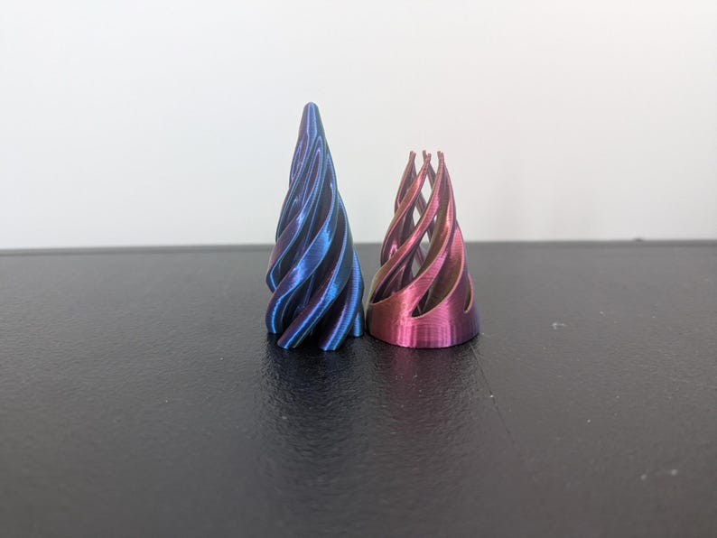 Może przedstawiać: Dwa stożkowe obiekty o spiralnej konstrukcji z otwartą ramą. Wyższy obiekt ma metaliczne wykończenie z gradientem niebiesko-fioletowym, a kr&oacute;tszy obiekt ma metaliczne wykończenie z gradientem r&oacute;żowo-fioletowym z widocznymi pionowymi żebrami.