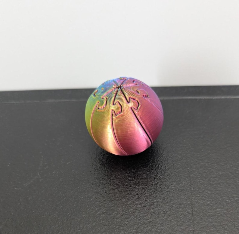 Op de afbeelding: Een bolvormig object met een regenboogverloop dat overgaat van blauw naar groen, geel en roze. Het oppervlak is voorzien van een reeks in elkaar grijpende, gebogen segmenten die aan de bovenkant samenkomen en een patroon van fijne verticale lijnen vormen.