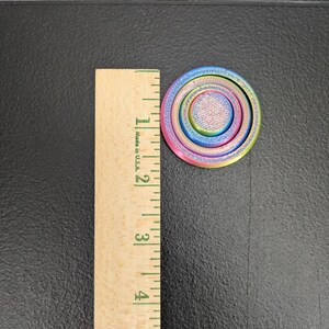 Peut inclure: Un fidget spinner circulaire multicolore avec un d&eacute;grad&eacute; de teintes bleues, violettes, roses et jaunes. L'objet est plac&eacute; &agrave; c&ocirc;t&eacute; d'une r&egrave;gle en bois, montrant un diam&egrave;tre d'environ 3,8 centim&egrave;tres pour ce dispositif de fidget circulaire.