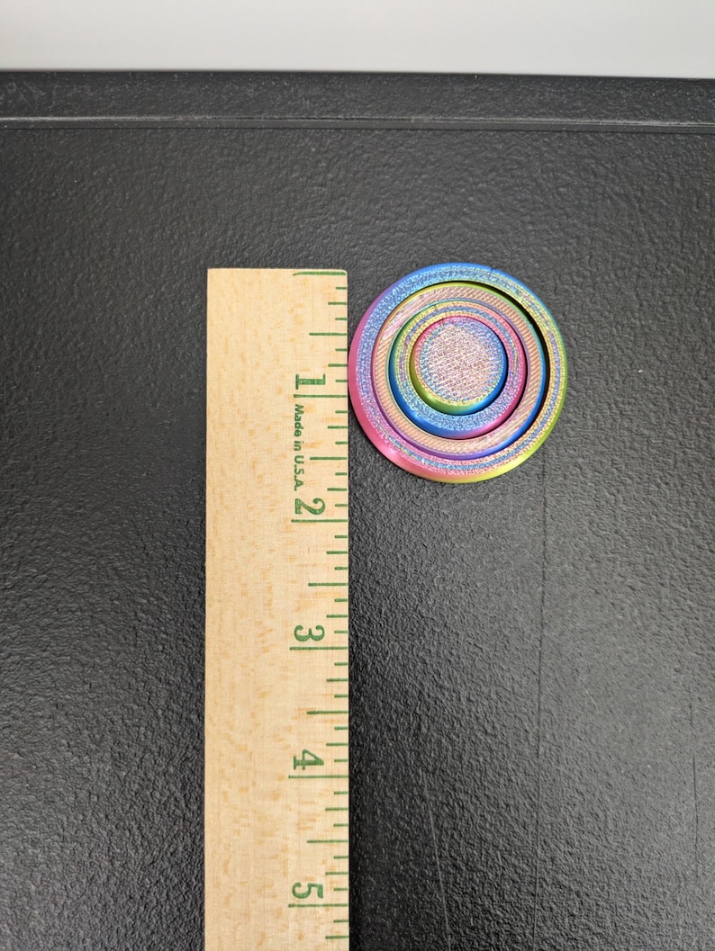 Peut inclure: Un fidget spinner circulaire multicolore avec un d&eacute;grad&eacute; de teintes bleues, violettes, roses et jaunes. L'objet est plac&eacute; &agrave; c&ocirc;t&eacute; d'une r&egrave;gle en bois, montrant un diam&egrave;tre d'environ 3,8 centim&egrave;tres pour ce dispositif de fidget circulaire.