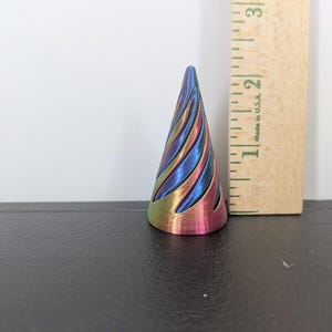 Może przedstawiać: Stożkowy przedmiot ze spiralnym wzorem wycięć, posiadający metaliczny tęczowy gradient w kolorach niebieskim, fioletowym i złotym. Drewniana linijka umieszczona obok przedmiotu wskazuje jego wysokość na około 5,7 centymetra.