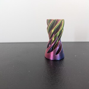 Może przedstawiać: Mały, cylindryczny przedmiot o spiralnym, skręconym wzorze. Powierzchnia ma metaliczne, opalizujące wykończenie, kt&oacute;re zmienia się między odcieniami fioletu, magenty, zieleni i złota, tworząc żywy, wielokolorowy wygląd.