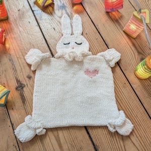 Peut inclure: Une couverture de sécurité tricotée de couleur crème représentant une tête de lapin avec des yeux fermés brodés et un petit nez rose. Le corps carré présente un col à volants, quatre coins à volants et un petit cœur rose sur le côté droit.