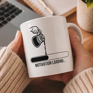 Op de afbeelding: Een witte keramische mok met een zwarte afbeelding van een hand die koffie uit een kan in een laadbalk giet. Onder de afbeelding staat de tekst MOTIVATION LOADING... in een vetgedrukt, schreefloos lettertype op de mok.