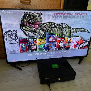 Peut inclure: Une console de jeu Xbox originale noire est placée devant un écran de télévision affichant un menu pour Jurassic Park: Operation Genesis. L'écran présente une grande illustration de dinosaure et une rangée de jaquettes de jeux Xbox.